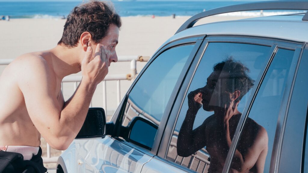 Man applying sunscreen to his face while looking in a car window at the beach